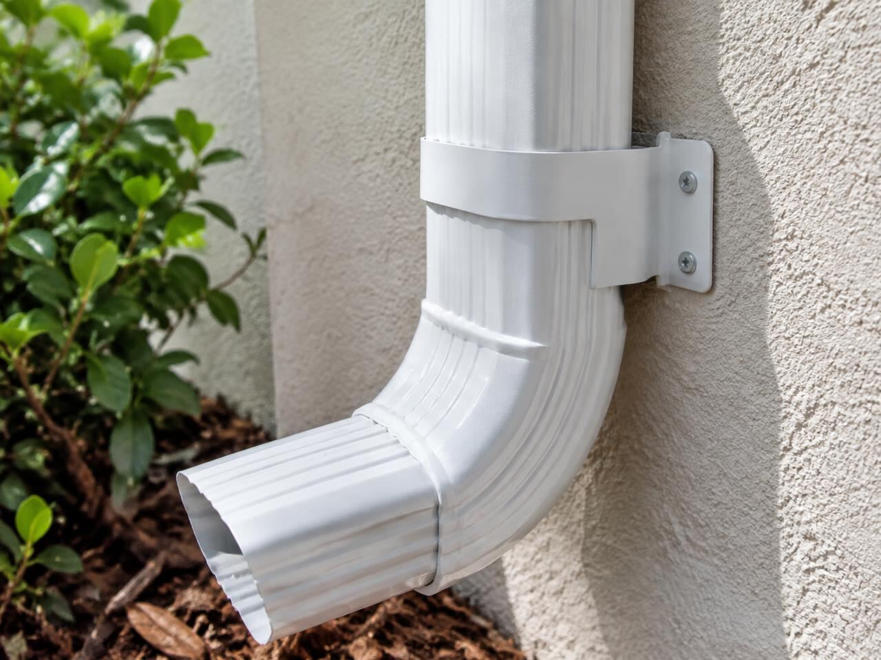 Close-up of a white aluminum downspout elbow and wall bracket on a stucco wall